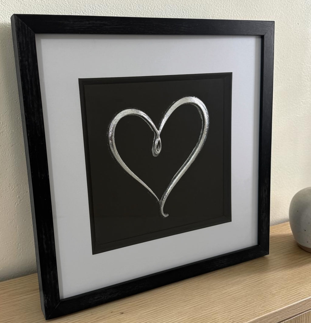 Silver love heart 3D relief artwork leaning on a wooden surface with black centre background and white mount.