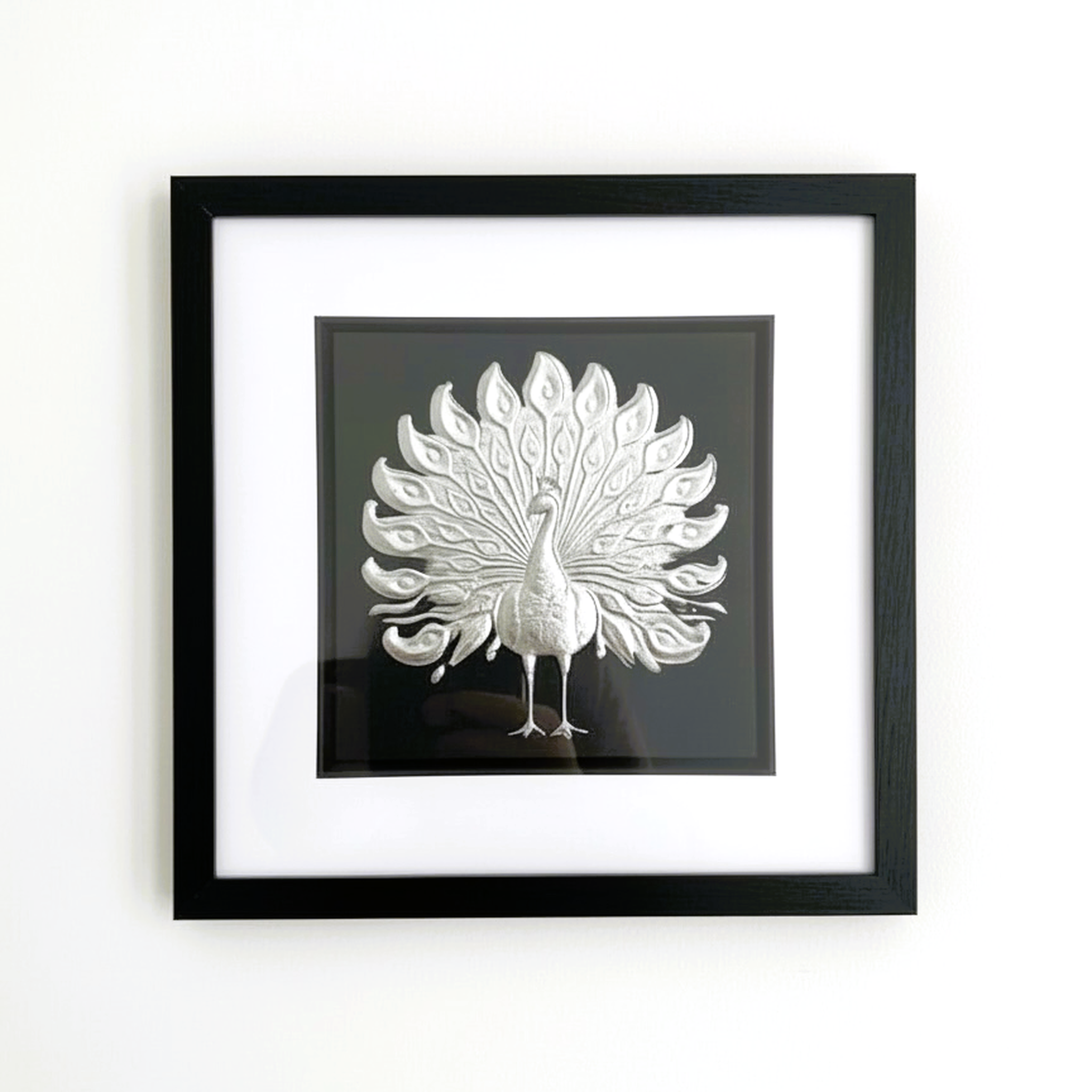Silver peacock wall art framed in black with white mount on a light wall