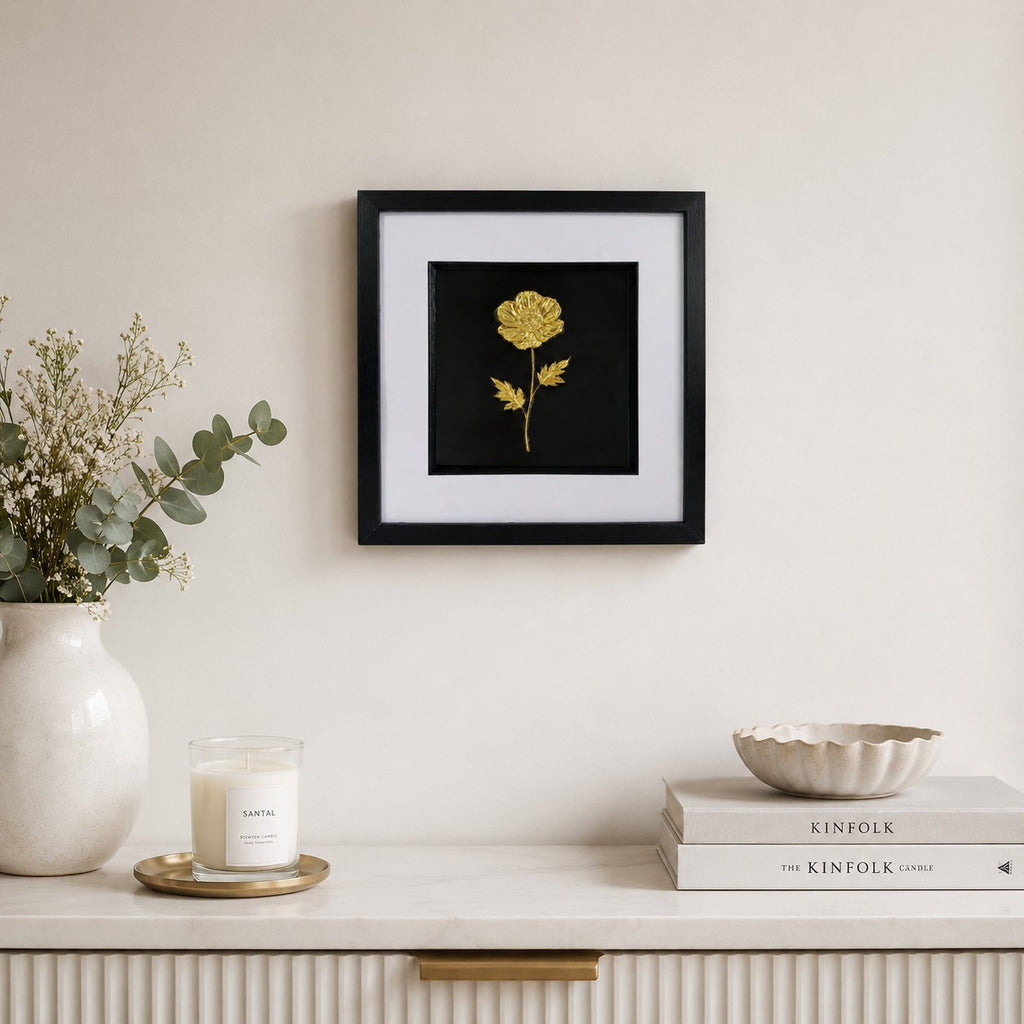 Gold framed wildflower artwork styled on a neutral wall above a pale sideboard with vase, candle and books.