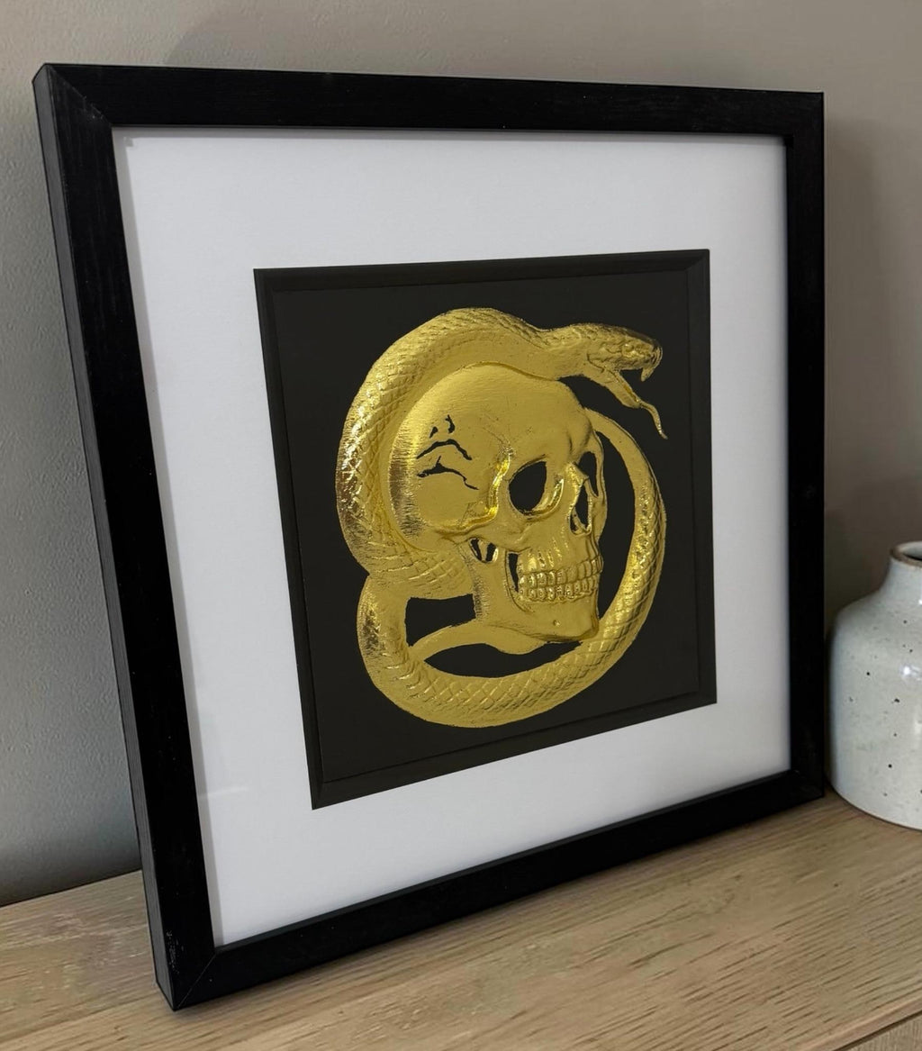 Gold skull and serpent relief artwork leaning on a wooden sideboard beside a small ceramic vase.