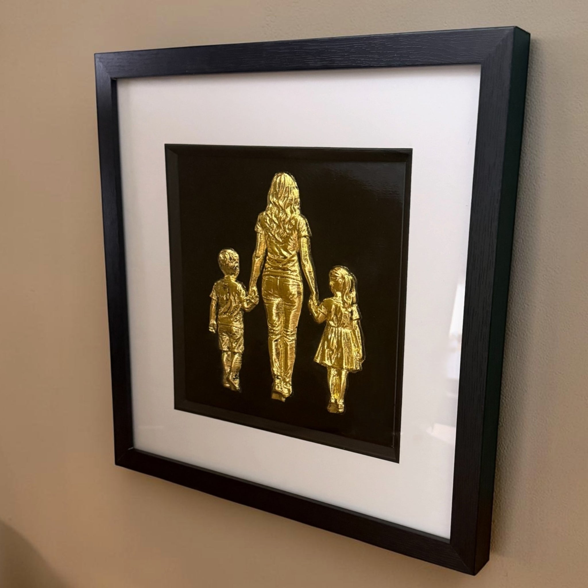 Mum, son and daughter framed wall art in gold leaf mounted on a wall, with black wooden frame, white mount and black background