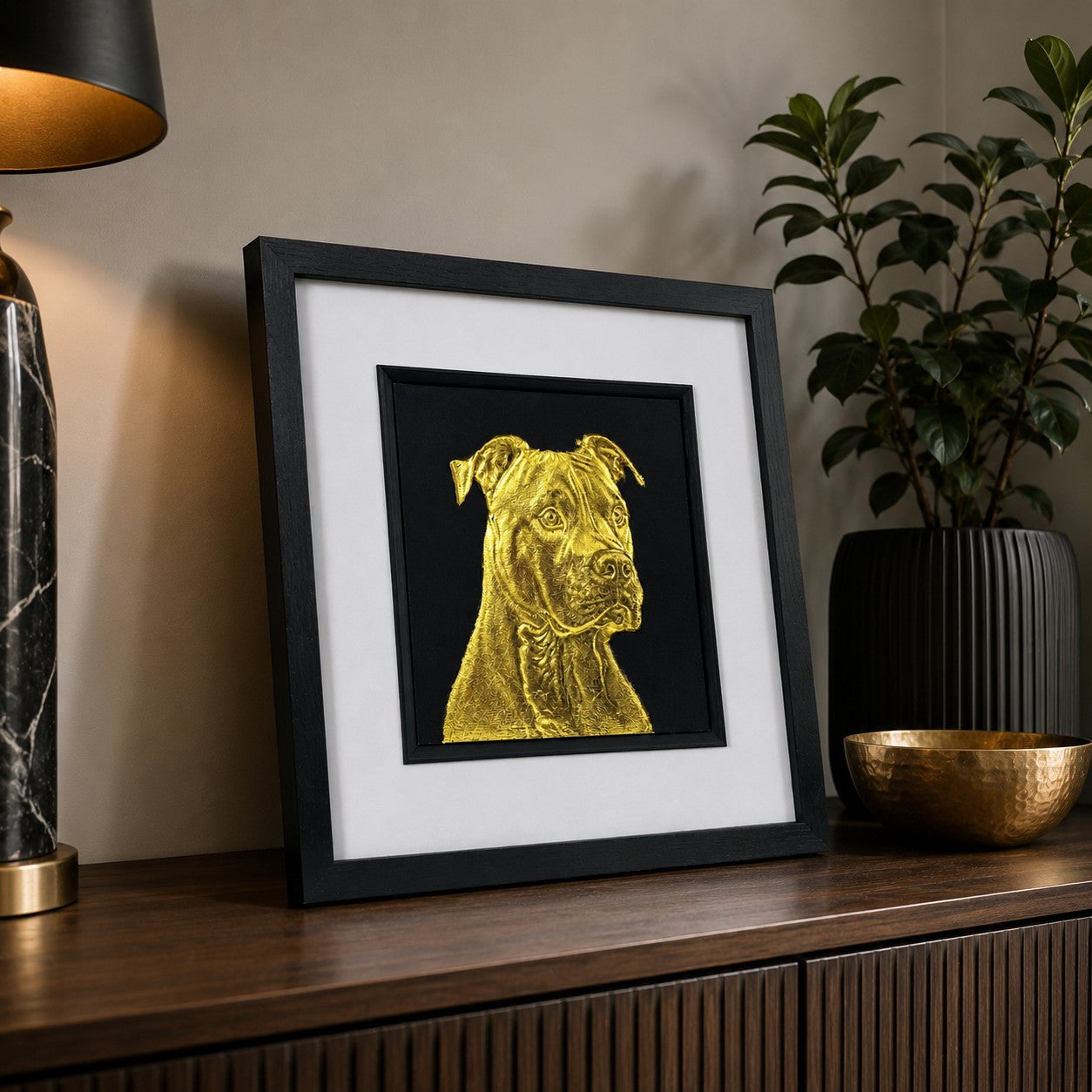 Angled lifestyle view of Staffordshire Bull Terrier gold leaf 3D wall art in a black frame, displayed with warm wood, black decor and brass accents in a modern home setting.