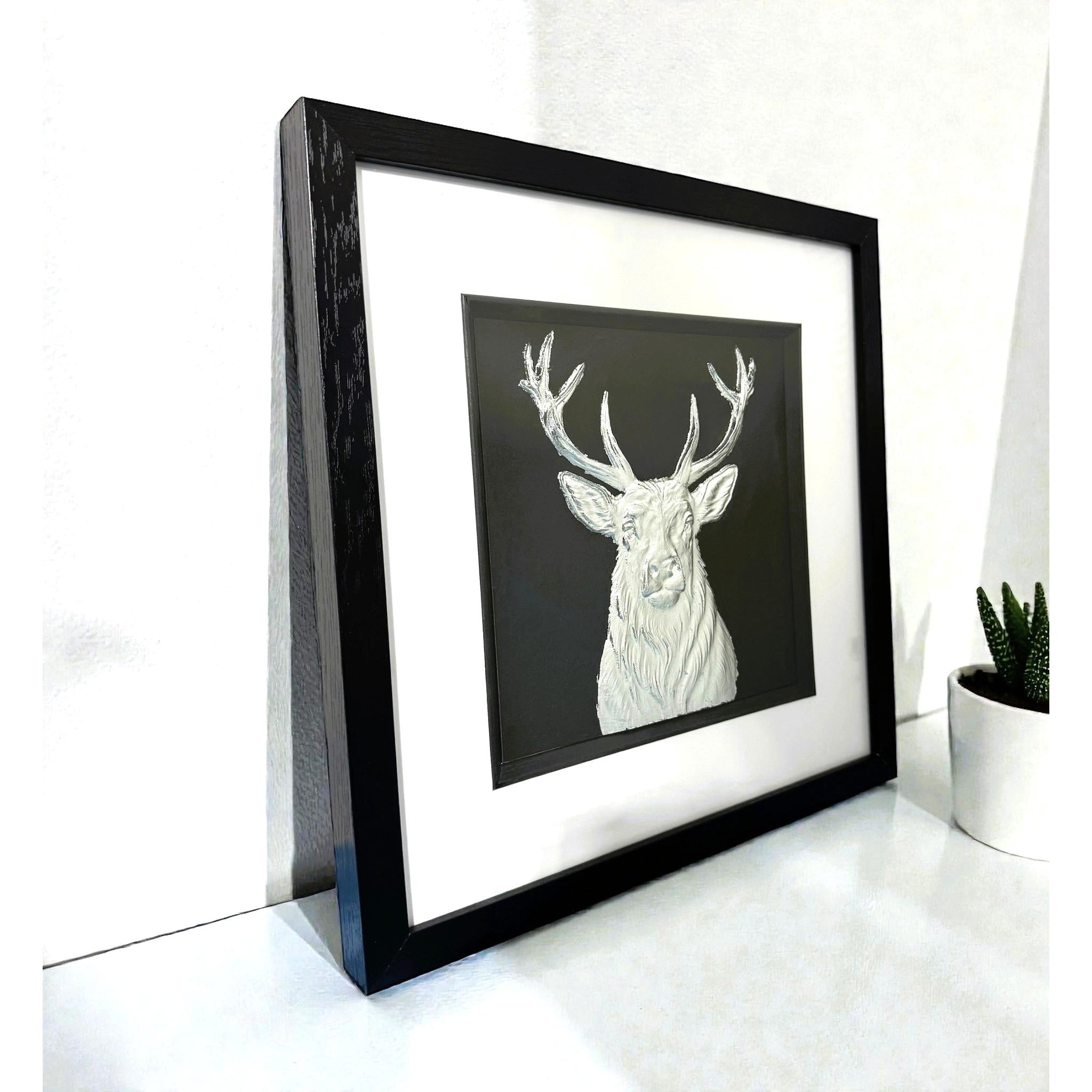 Silver stag wall art framed in black with white mount displayed on a shelf