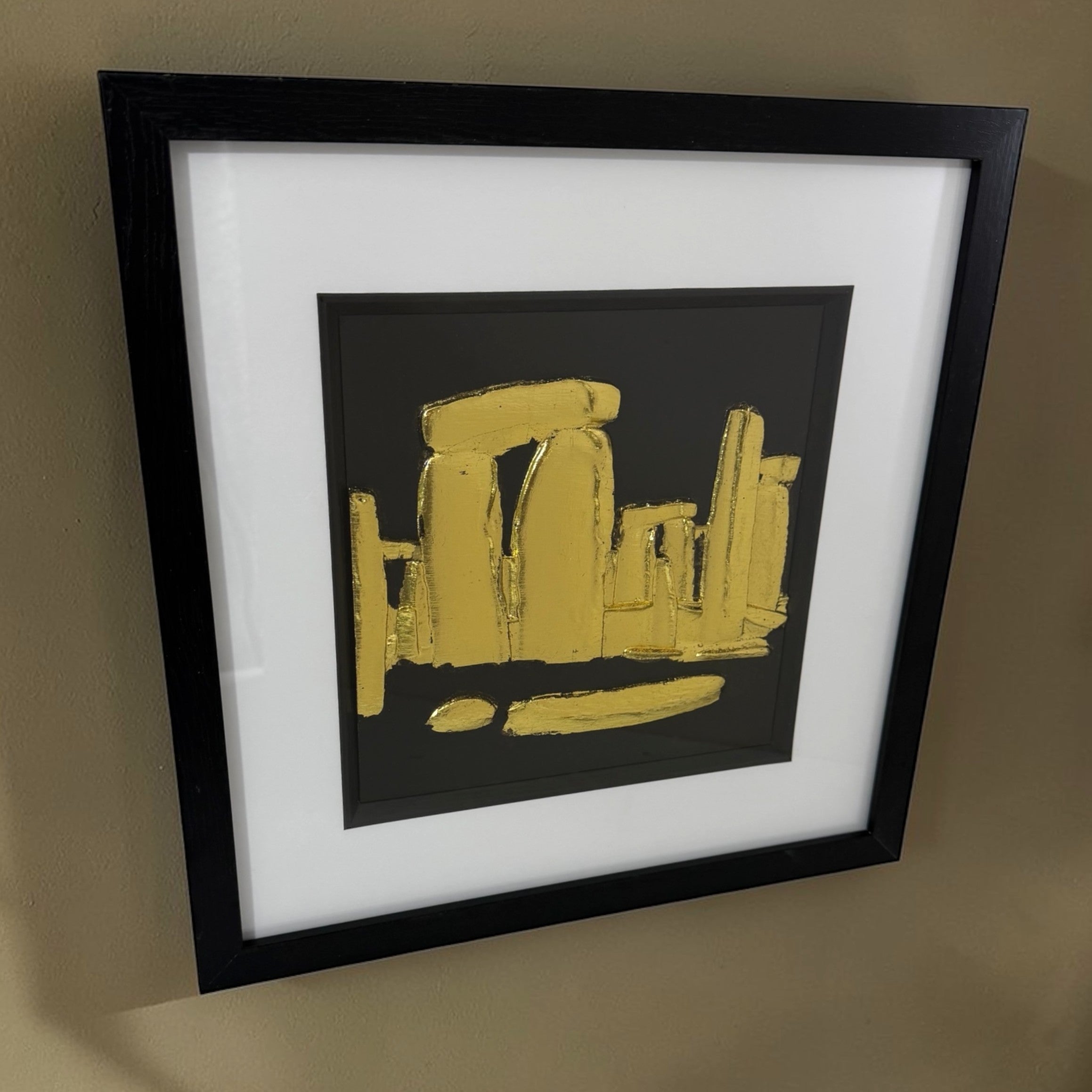 Angled view of gold Stonehenge raised relief wall decor in a black frame on a neutral wall.