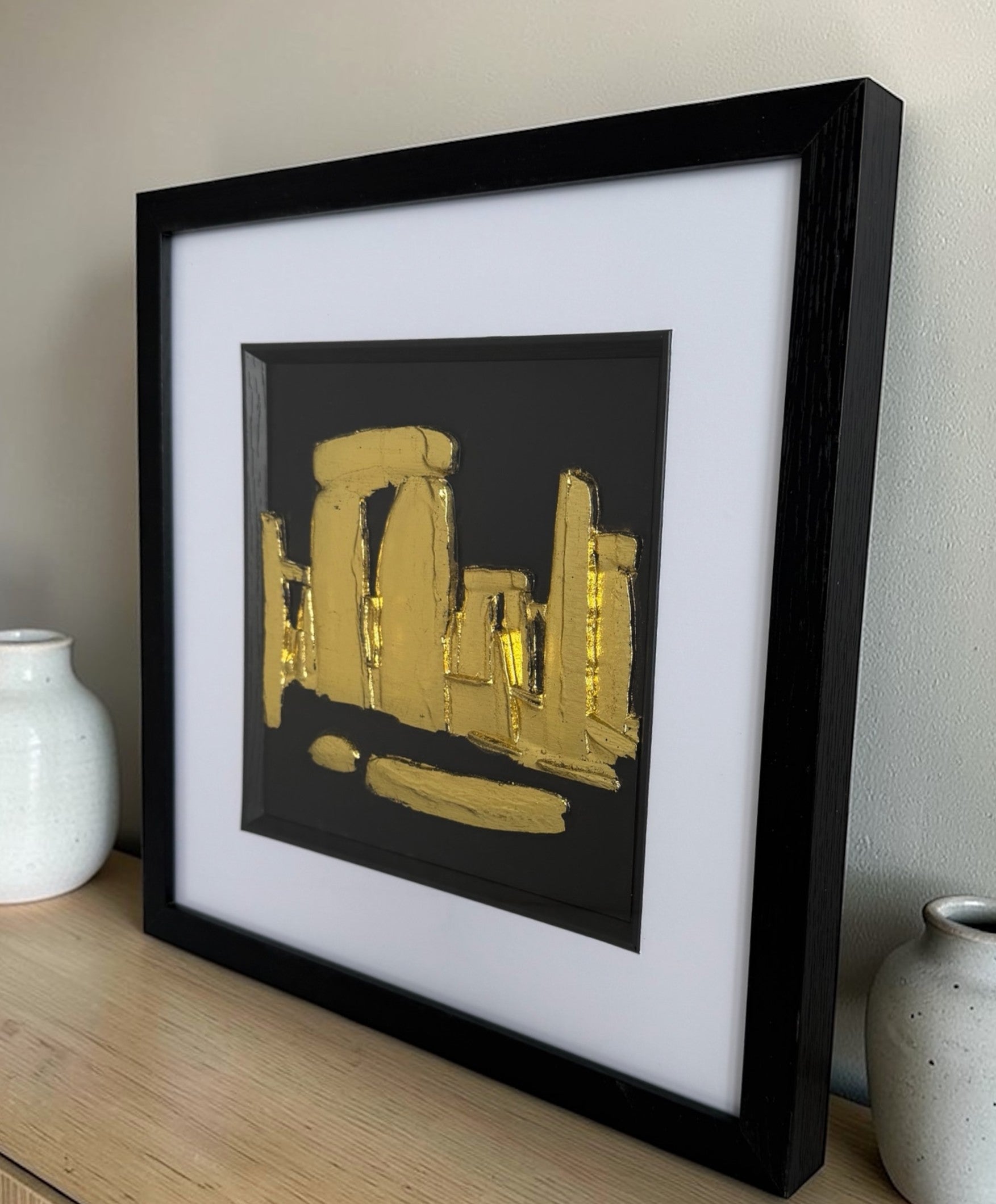 Gold Stonehenge raised relief in a black frame with white mount and glass front, styled on a wooden sideboard beside ceramic vases.