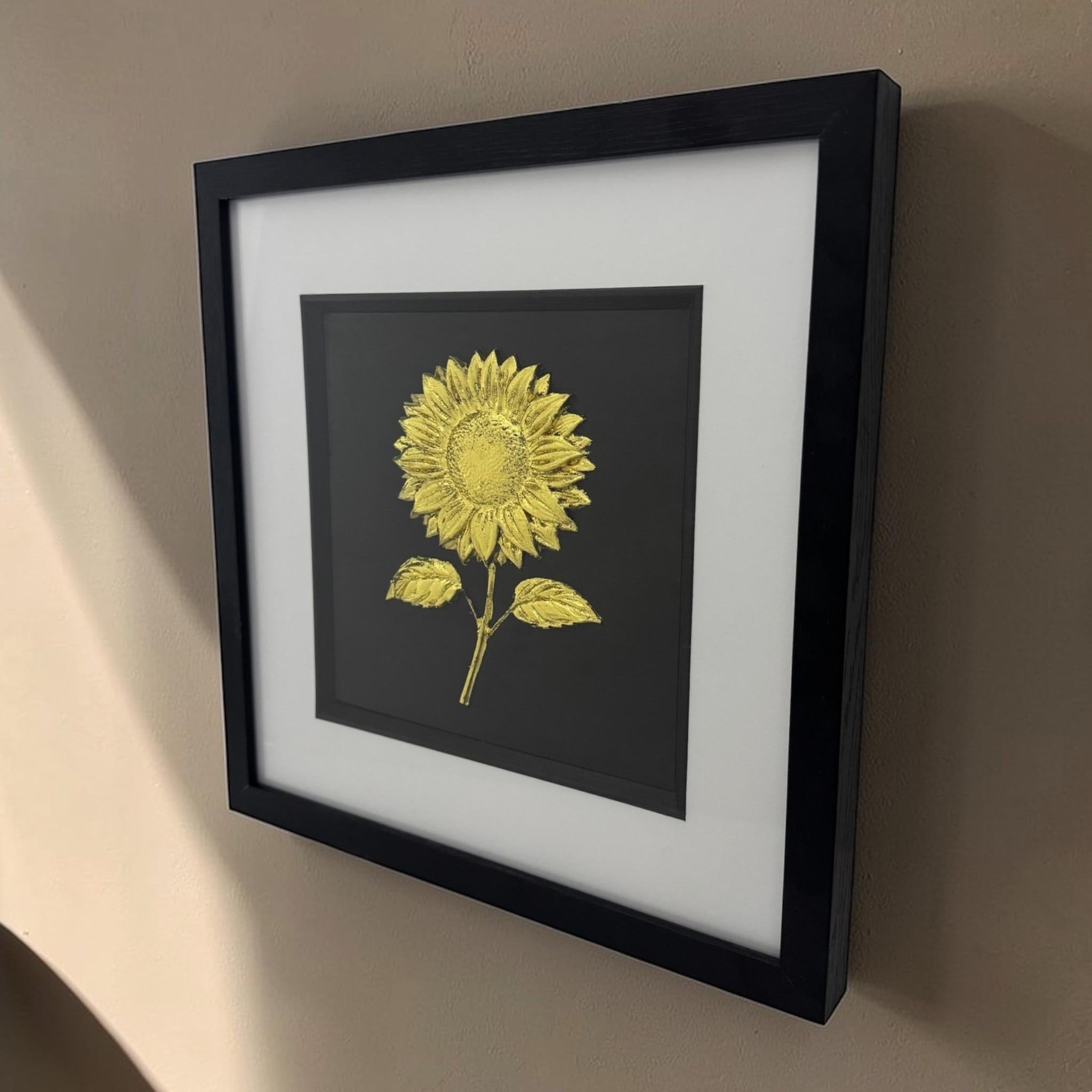 Gold Leaf sunflower relief artwork hung on a neutral wall, angled view showing the depth of the black box frame and white mount
