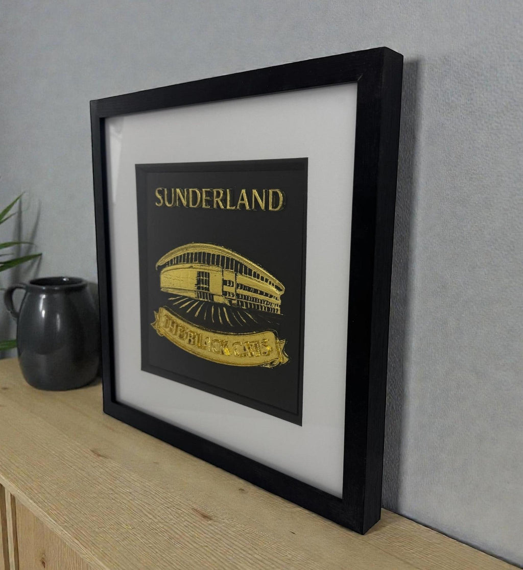 Sunderland framed wall art in gold leaf 3D relief, deep black box frame with white mount, styled on a wooden surface against a grey wall.