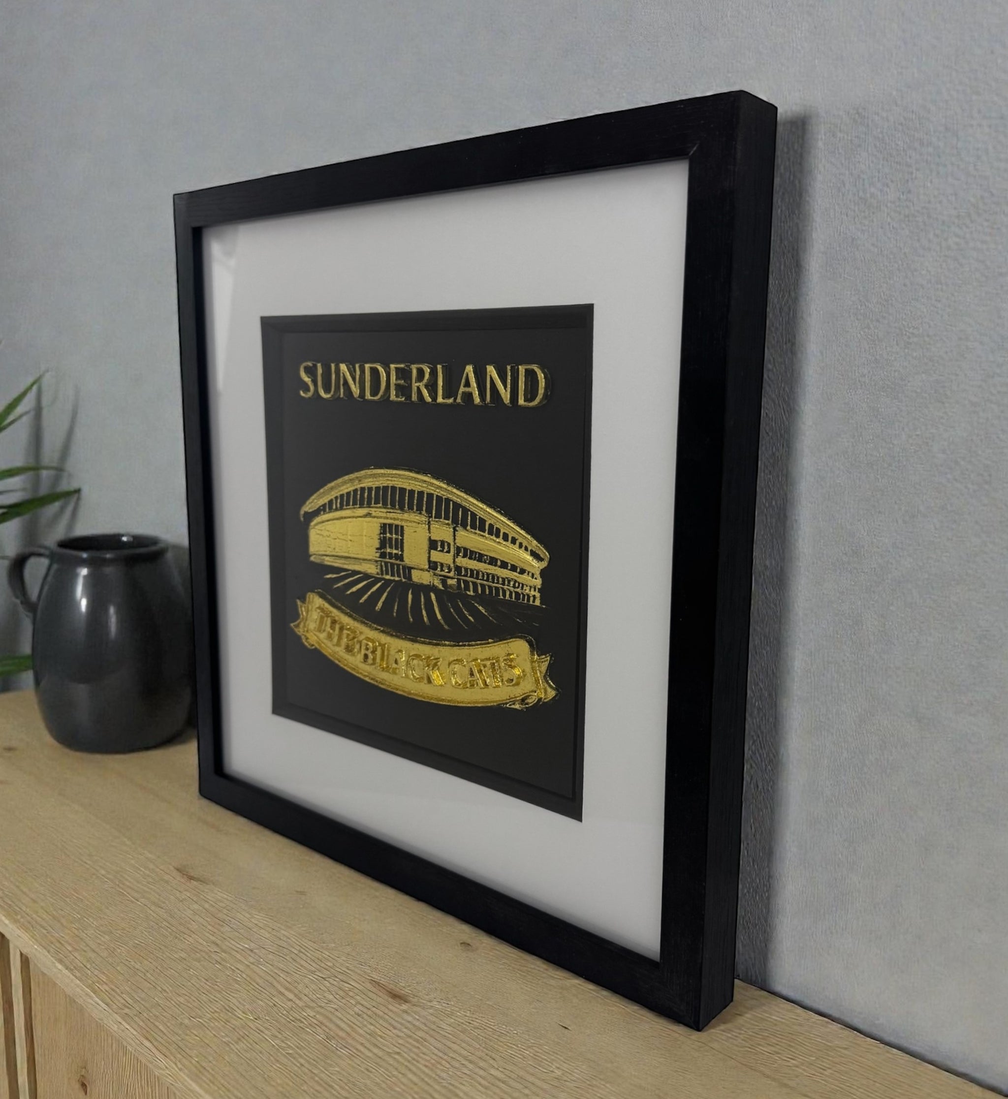 Sunderland framed wall art in gold leaf 3D relief, deep black box frame with white mount, styled on a wooden surface against a grey wall.