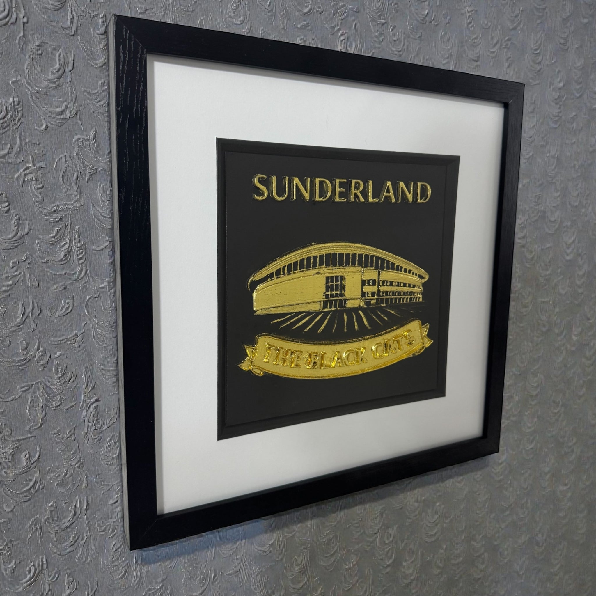 Sunderland 3D wall art in gold leaf relief, black box frame with white mount, shown hanging on a textured grey wall at an angle.