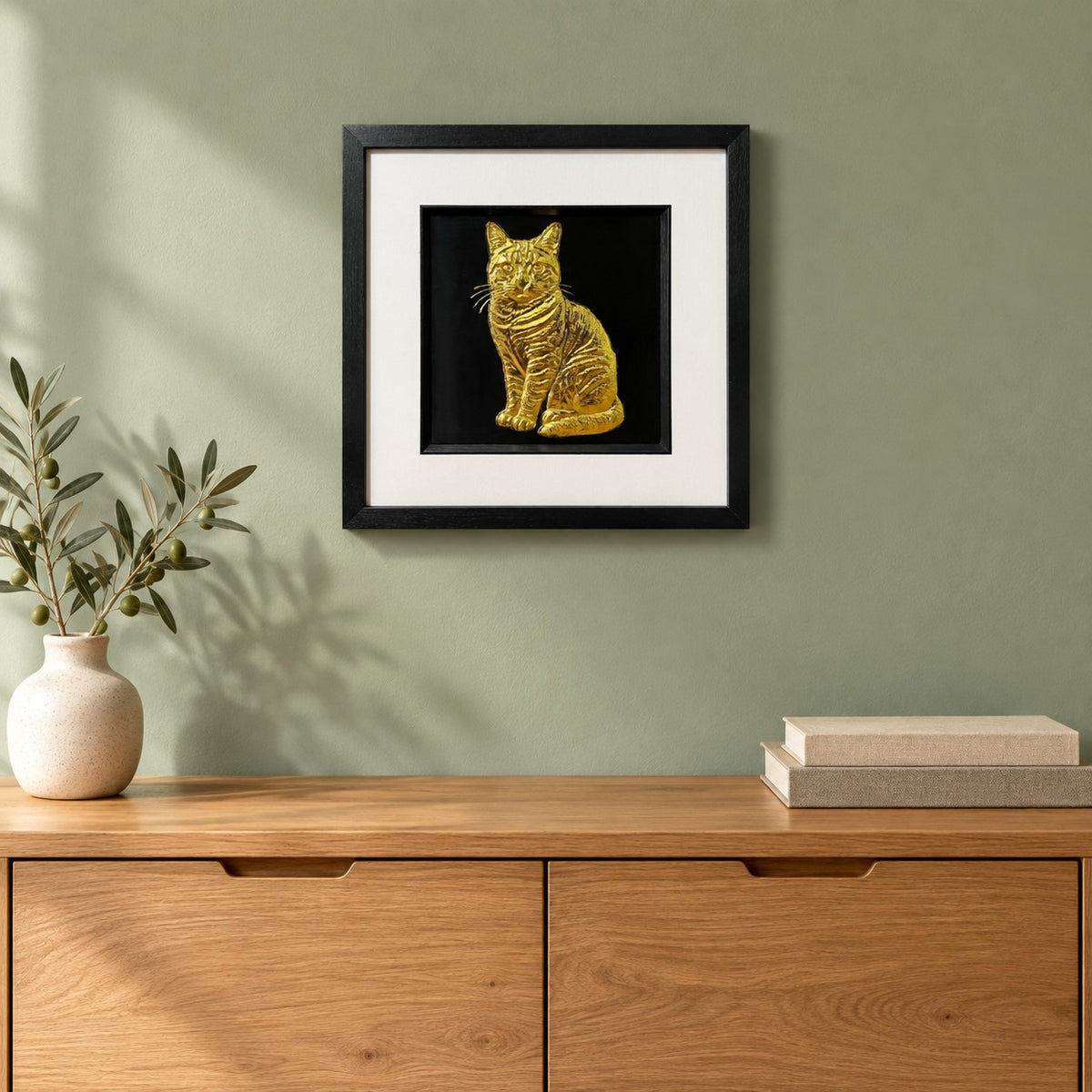 Gold tabby cat 3D wall art in a black square frame hung on a sage green wall above an oak sideboard with a vase of olive branches and two linen books
