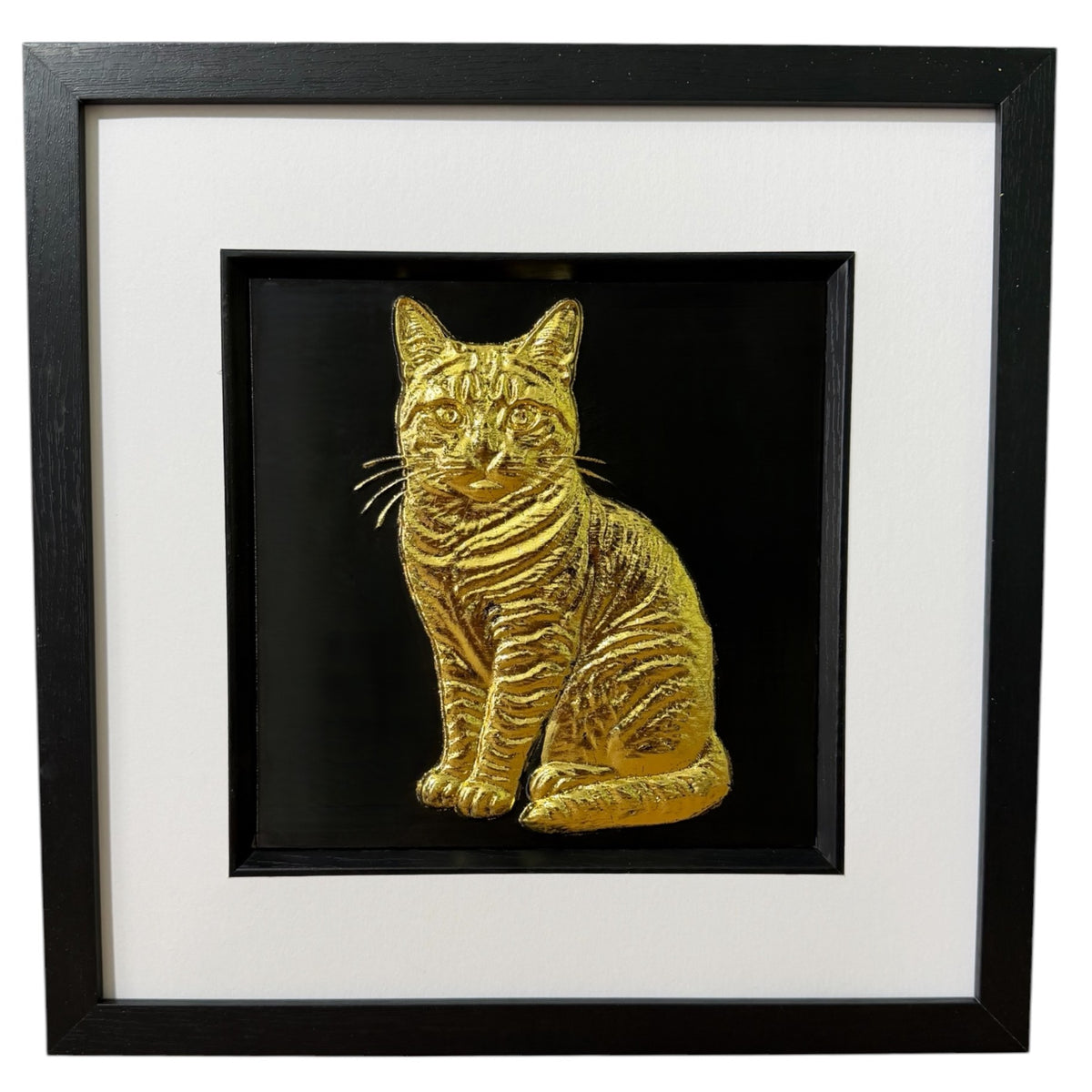 Framed tabby cat wall art, gold relief of a sitting striped tabby, tail curled round its paws, on a black background in a black wooden frame with white mount