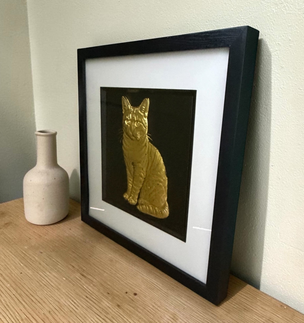 Gold tabby cat framed wall art leaning on an oak sideboard beside a cream bottle vase, angled to show the raised relief stripes through the glass