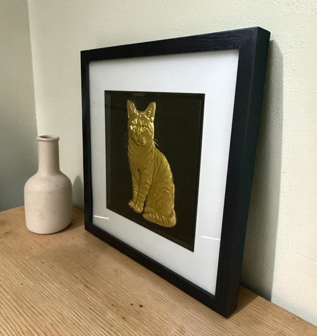 Gold tabby cat framed wall art leaning on an oak sideboard beside a cream bottle vase, angled to show the raised relief stripes through the glass