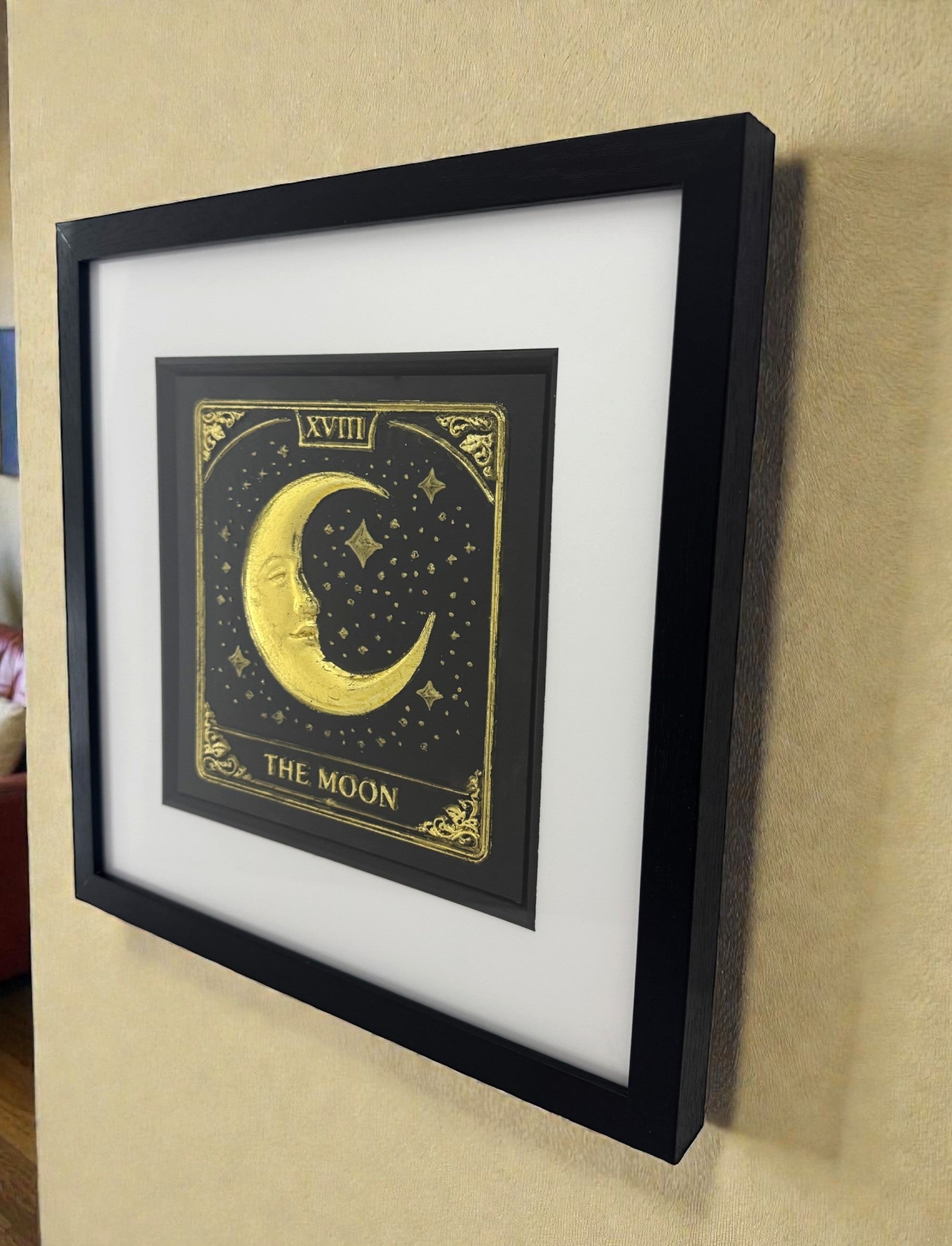 The Moon tarot card wall art in gold leaf 3D relief, framed in black with white mount, angled wall view showing depth and shadows