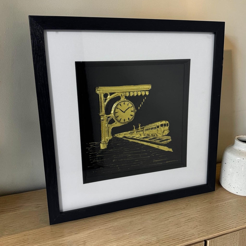 Gold train scene with station clock styled on a wooden sideboard in a black box frame beside a ceramic vase.