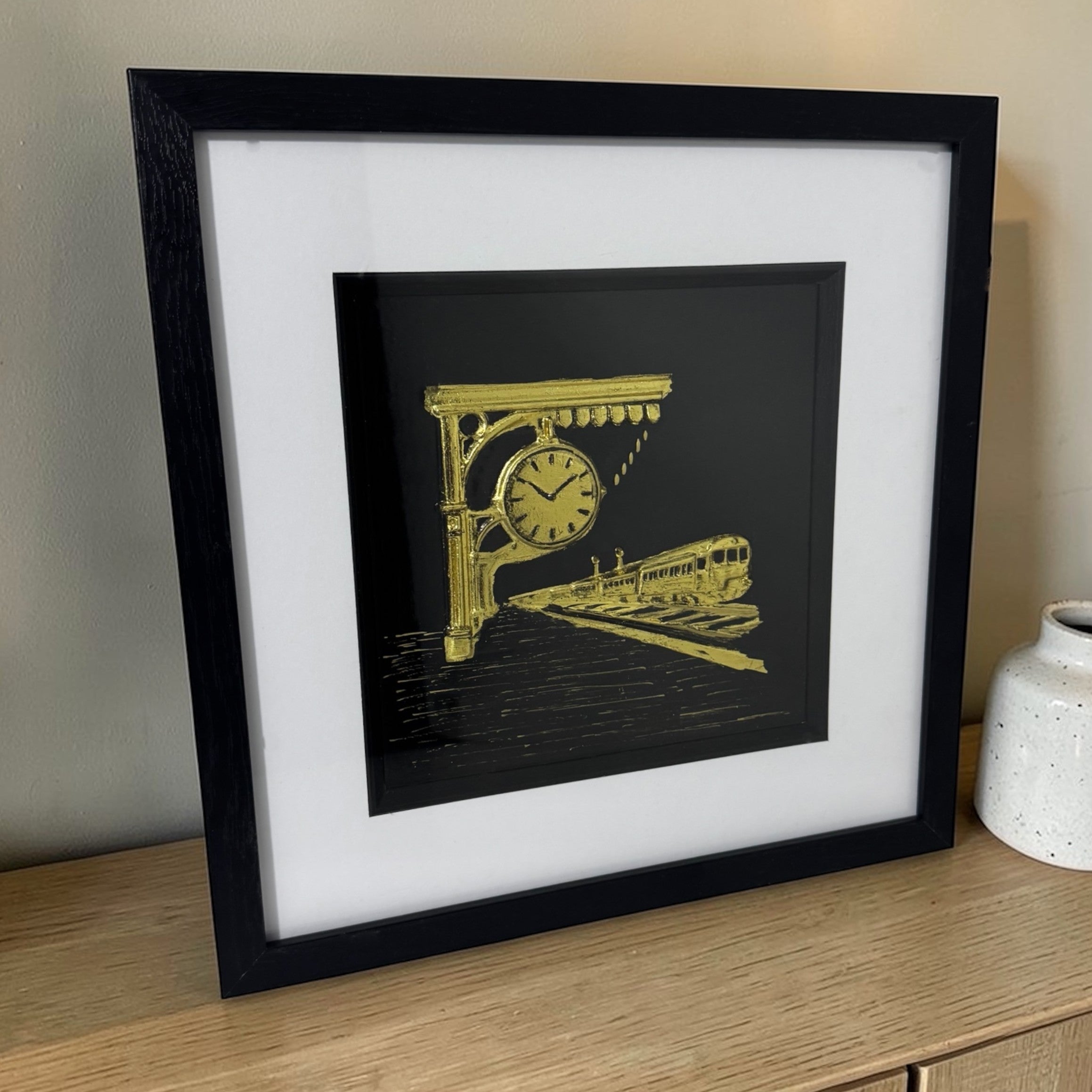 Gold train scene with station clock styled on a wooden sideboard in a black box frame beside a ceramic vase.