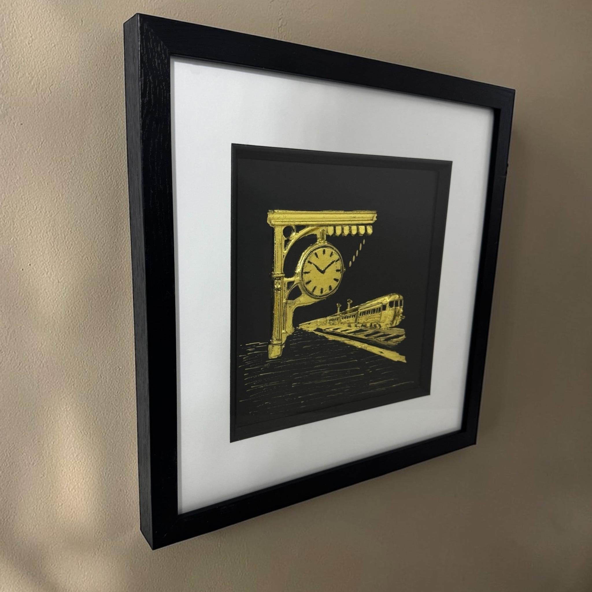 Angled view of a gold train station relief in a black box frame hanging on a neutral wall.
