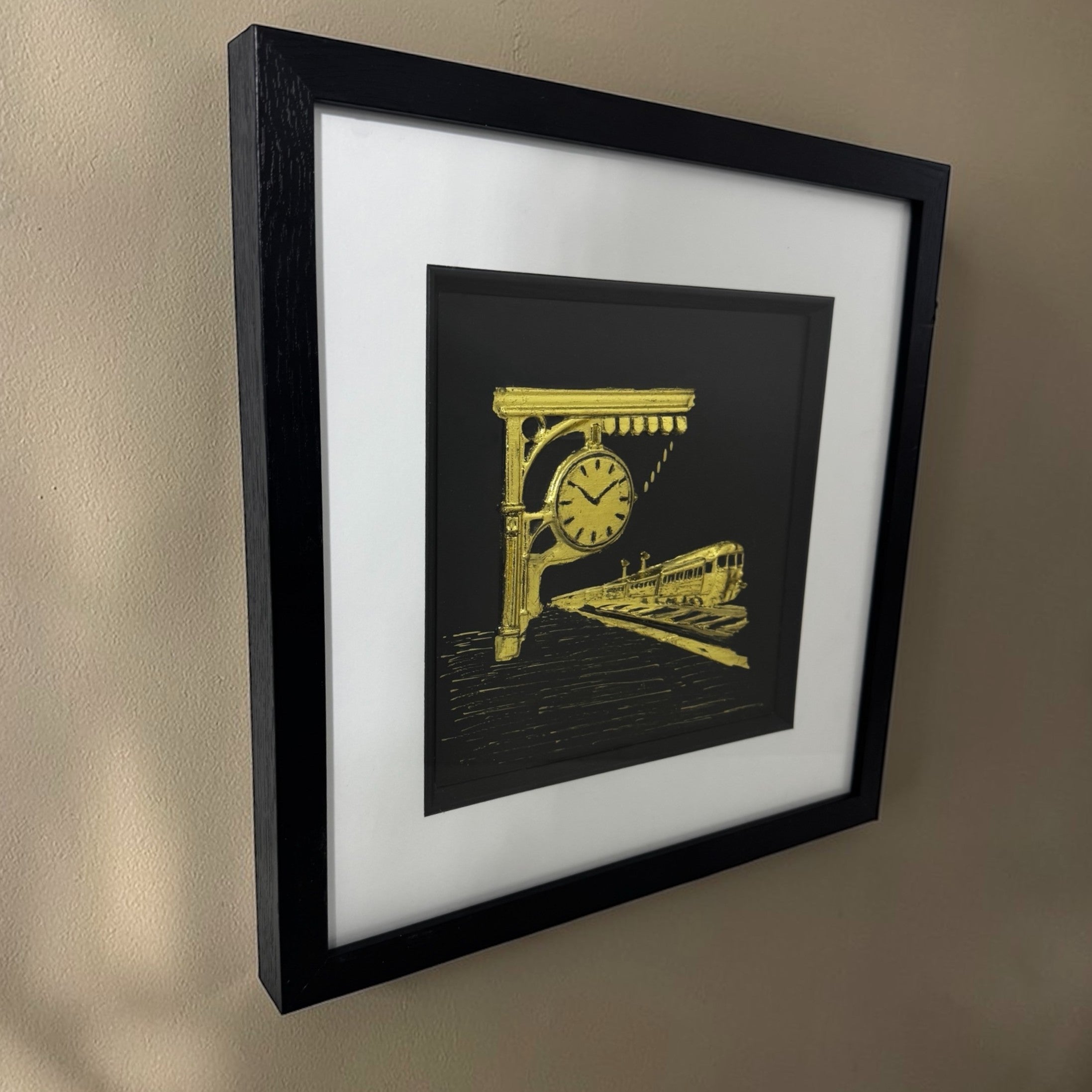 Angled view of a gold train station relief in a black box frame hanging on a neutral wall.