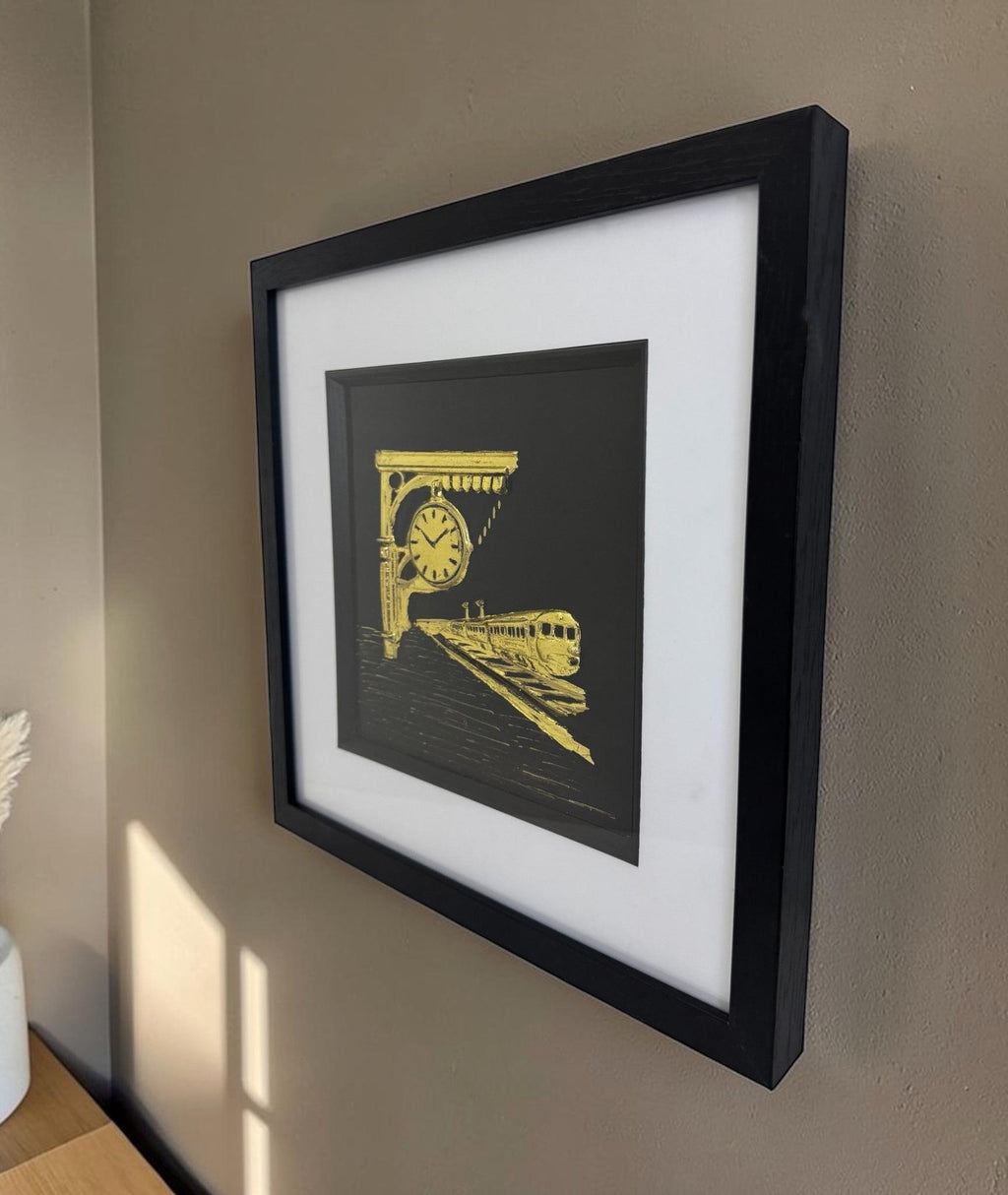 Angled side view of a gold train station relief in a black box frame hanging on a neutral wall. Showing the relefi depth