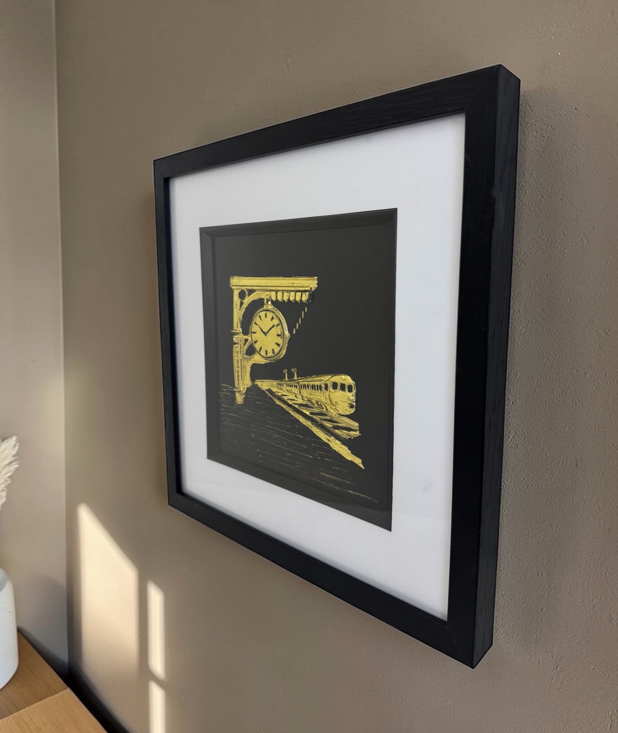 Angled side view of a gold train station relief in a black box frame hanging on a neutral wall. Showing the relefi depth