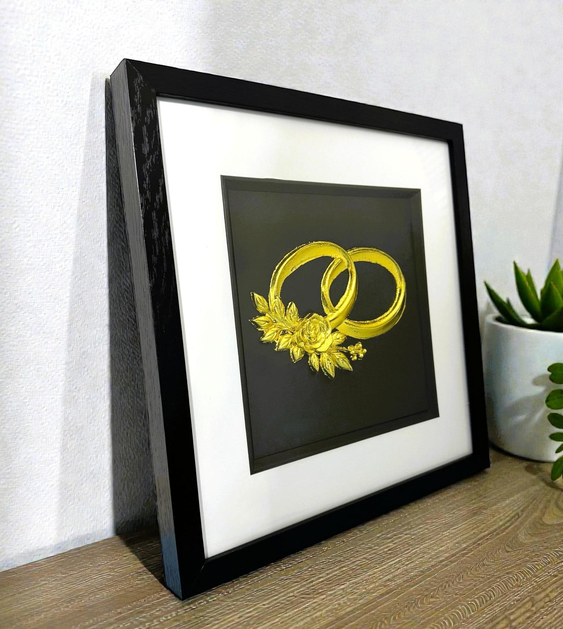 Angled desk view of gold leaf wedding rings wall art on a black background in a black wooden frame