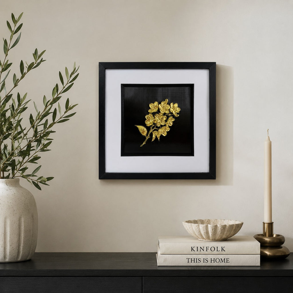Gold 3D flower bouquet artwork in a black box frame styled above a black sideboard with books, candle and olive branches