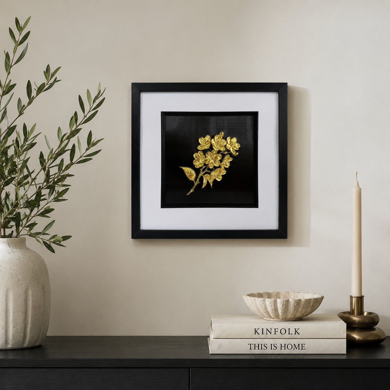 Gold 3D flower bouquet artwork in a black box frame styled above a black sideboard with books, candle and olive branches