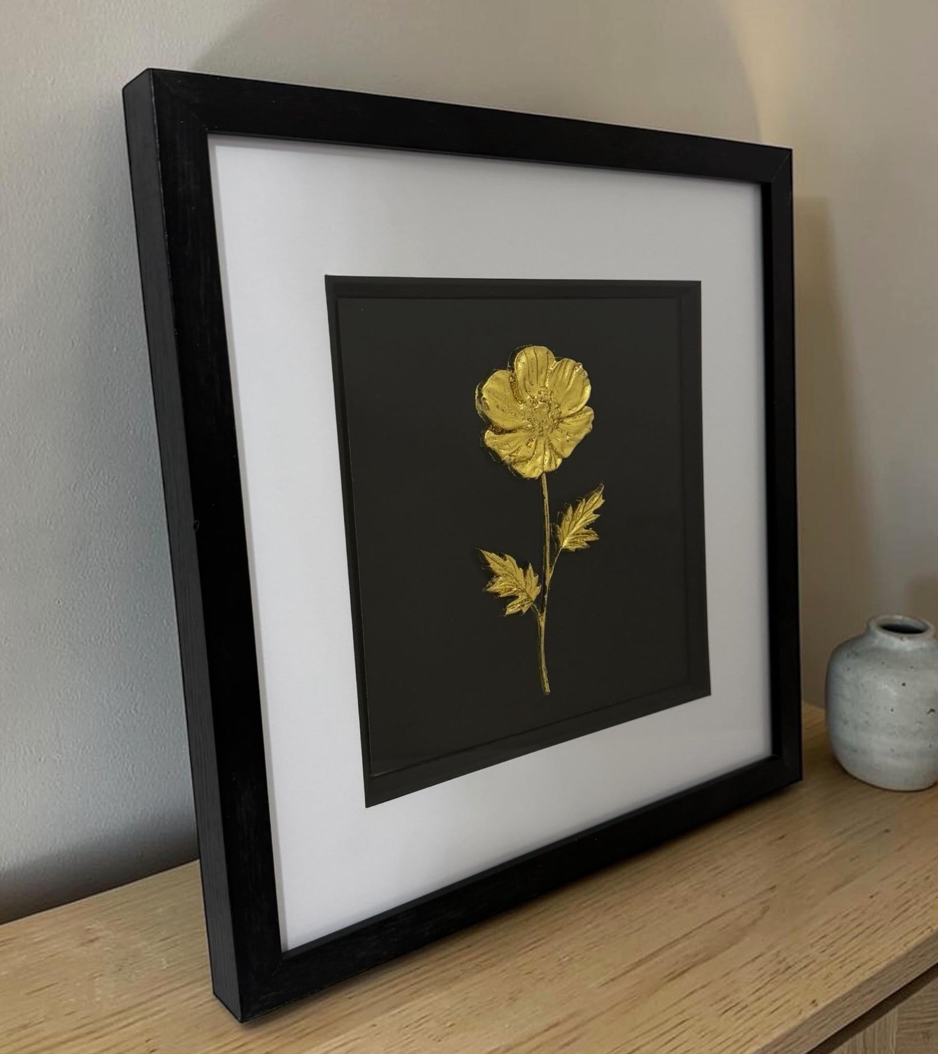 Angled gold wildflower artwork on a wooden sideboard showing black box frame depth and glass front.