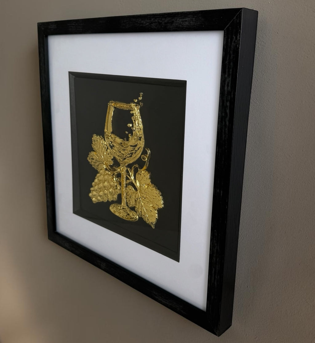 Gold wine glass and grape framed artwork angled on a wall showing square frame and layered mount.