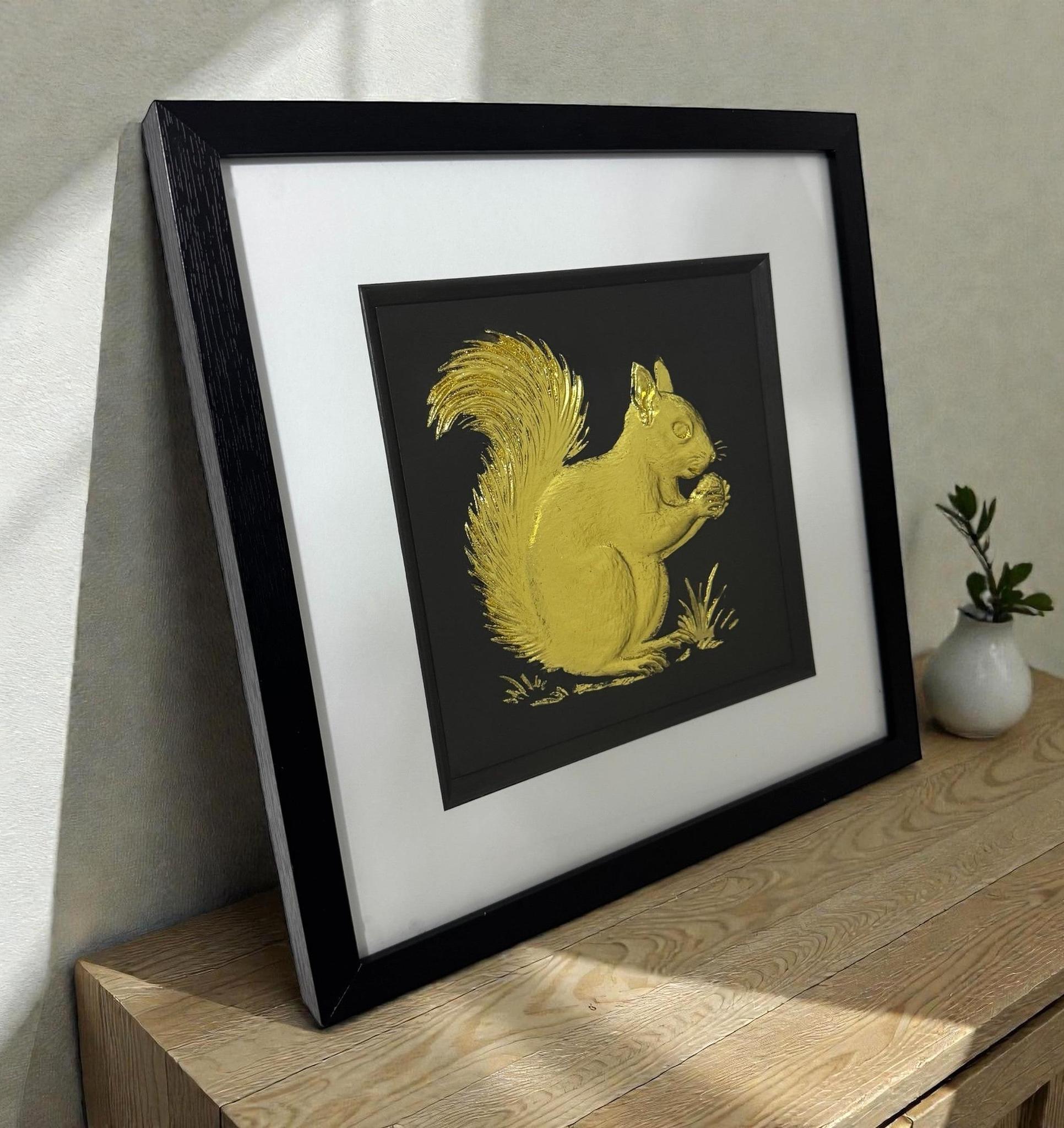 Framed red squirrel 3D wall art in gold leaf, in a black frame with white mount, styled on a wooden surface in a modern room.
