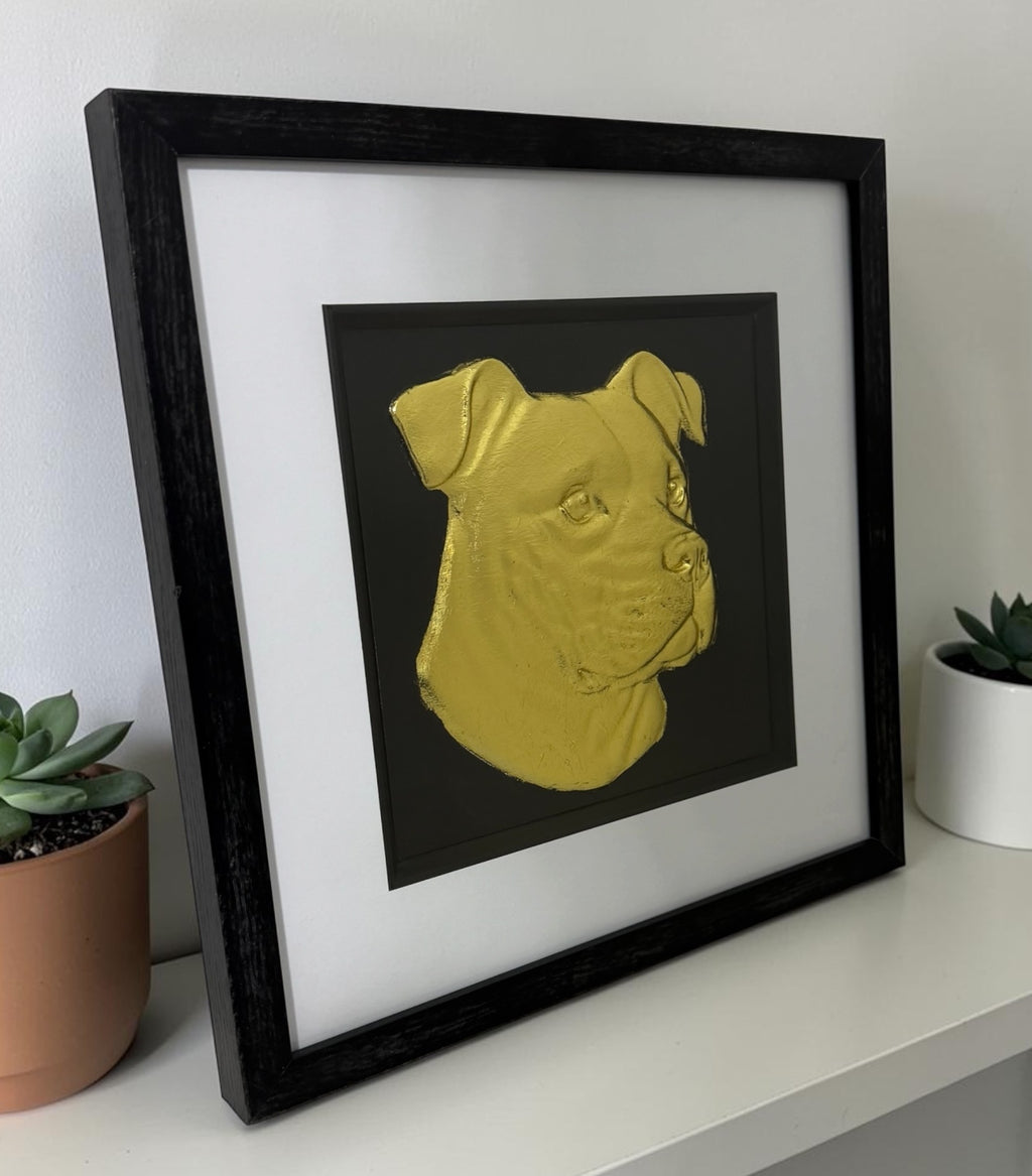 Angled lifestyle view of framed XL Bully dog wall art in gold leaf, displayed with a black frame and white mount beside small plants on a clean shelf.