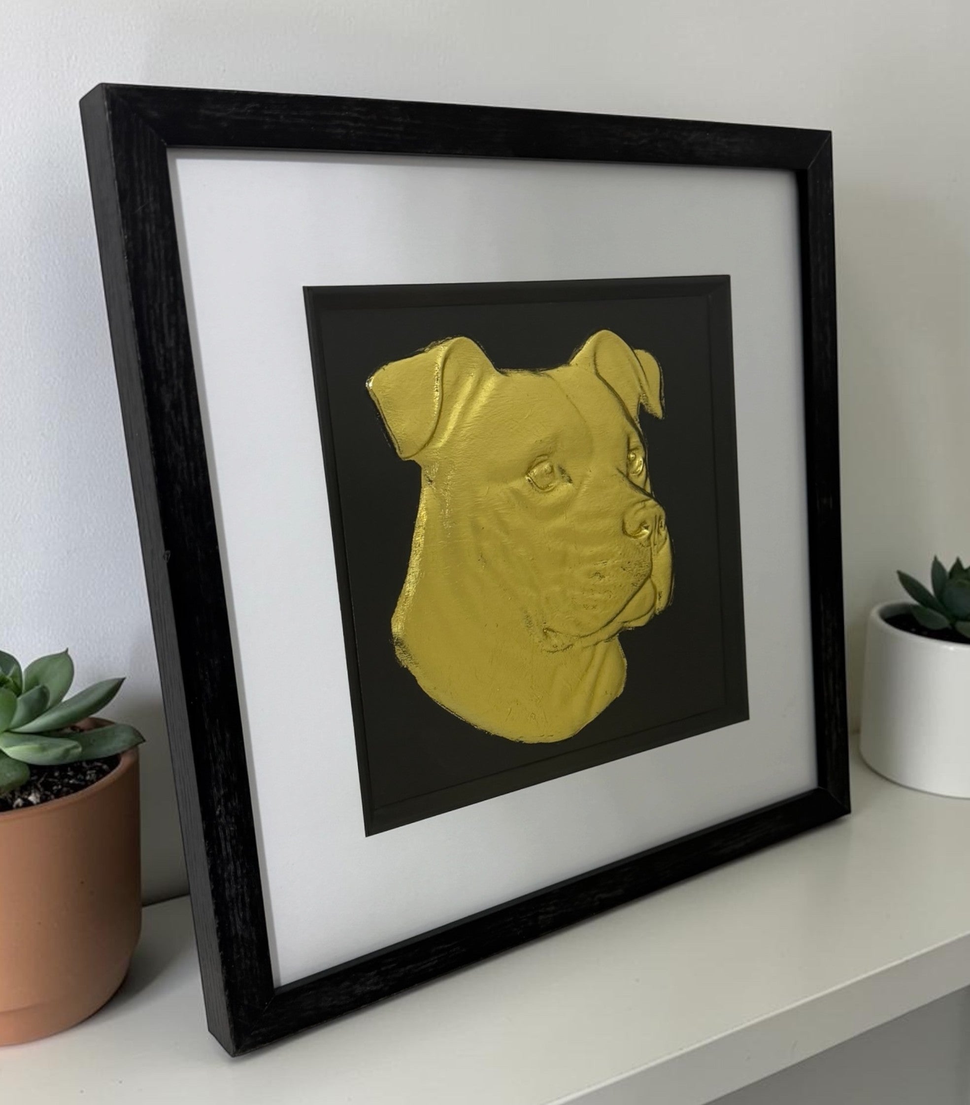 Angled lifestyle view of framed XL Bully dog wall art in gold leaf, displayed with a black frame and white mount beside small plants on a clean shelf.